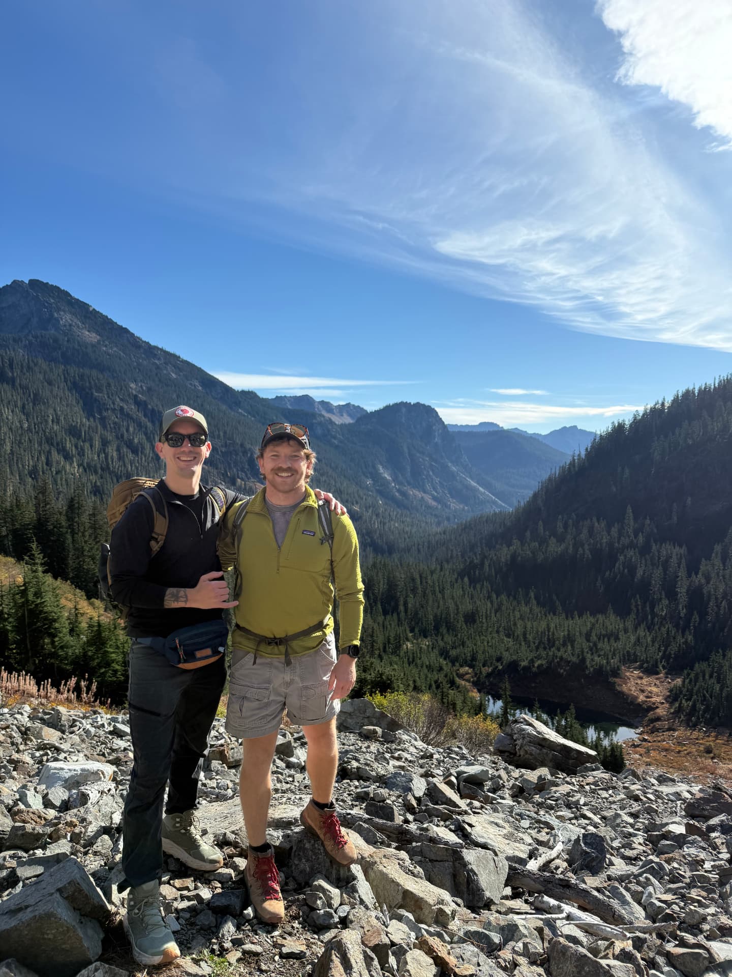 Two hikers stand on the rocky Source Lake overlook with the lake and forested valley above Alpental behind them